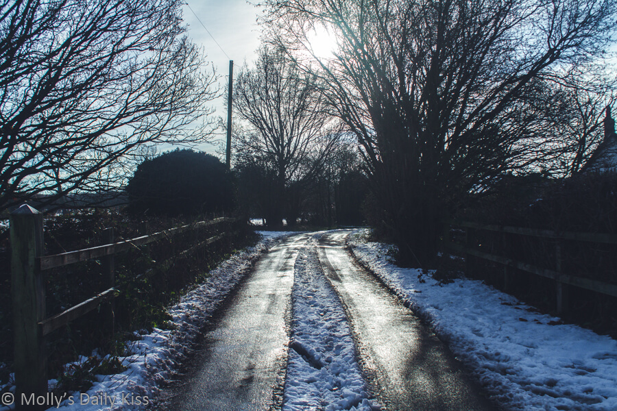 Snow lining either side of country lane with weak sunshine coming through the trees