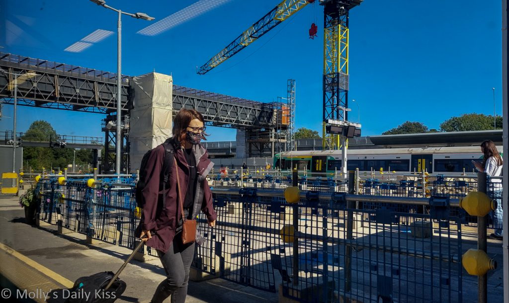 Person with face mask on at train station with construction going on round them and blue skies